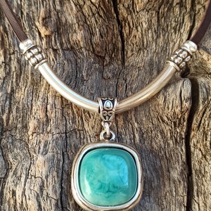 May include: A silver necklace with a brown leather cord and a square pendant featuring a teal green stone. The pendant has a small pearl hanging from the bottom.