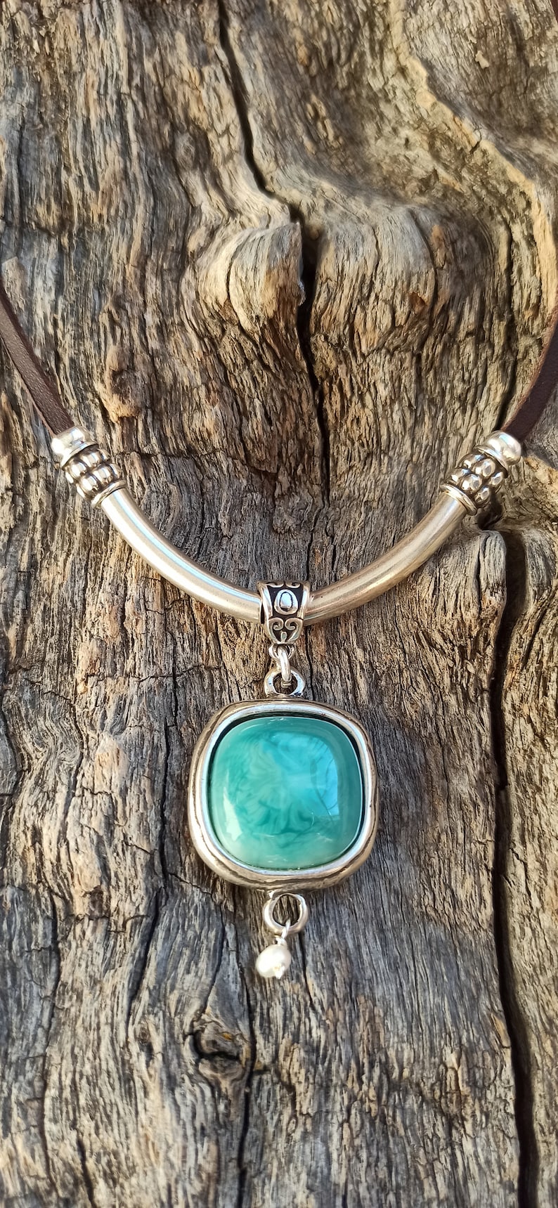 May include: A silver necklace with a brown leather cord and a square pendant featuring a teal green stone. The pendant has a small pearl hanging from the bottom.