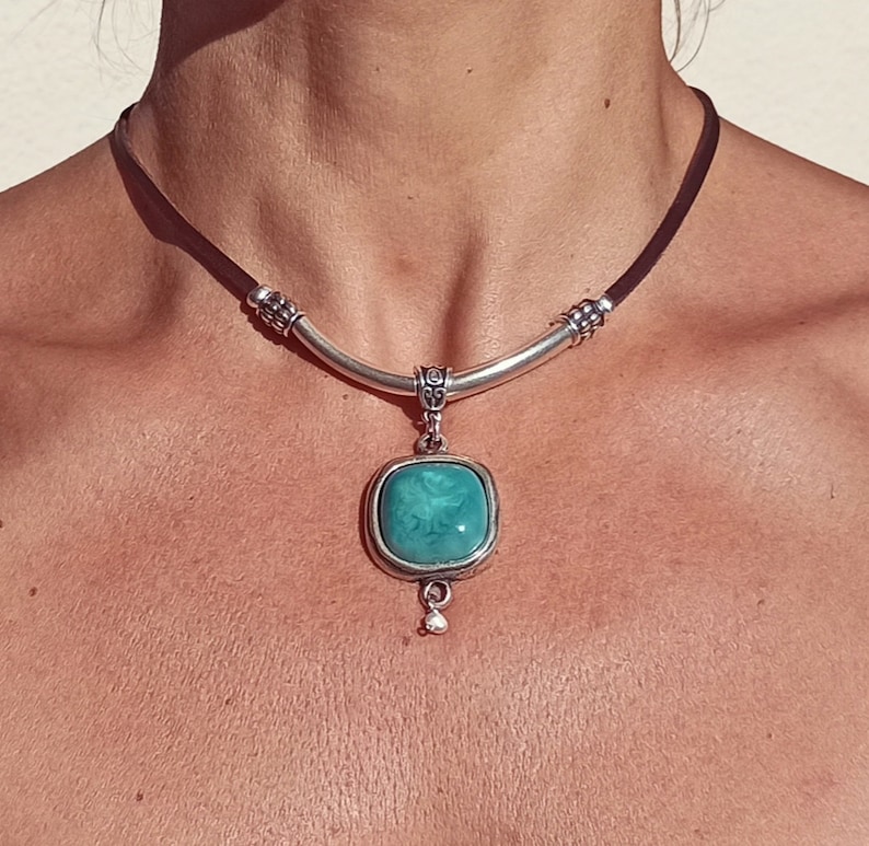 May include: A silver necklace with a brown leather cord and a turquoise pendant. The pendant is a square with rounded corners and has a silver frame. The pendant has a small silver bead hanging from the bottom.