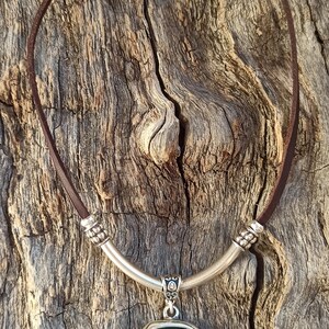 May include: A brown leather necklace with a silver pendant featuring a turquoise-colored stone. The pendant has a decorative design and hangs from a silver bar.