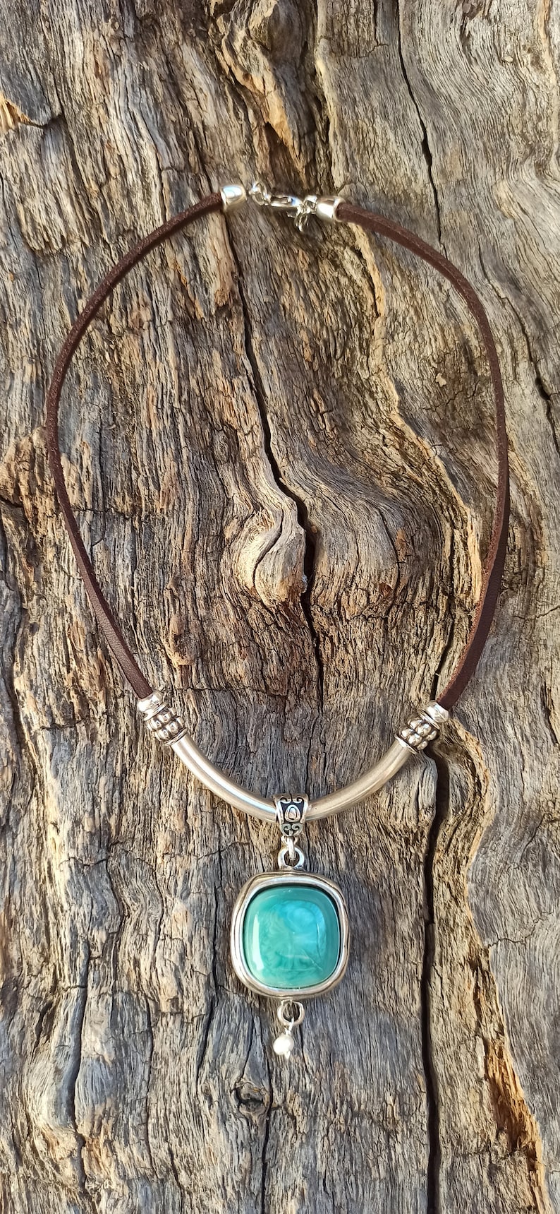 May include: A brown leather necklace with a silver pendant featuring a turquoise-colored stone. The pendant has a decorative design and hangs from a silver bar.