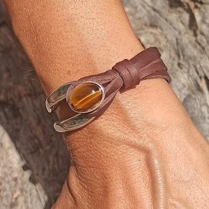 Op de afbeelding: Een armband van bruin leren koord met een zilverkleurige metalen sluiting in de vorm van een hoefijzer. Het midden van de sluiting is bezet met een ovale, gladde, goudbruine tijgeroog-edelsteen met een duidelijke chatoyante lichtband.