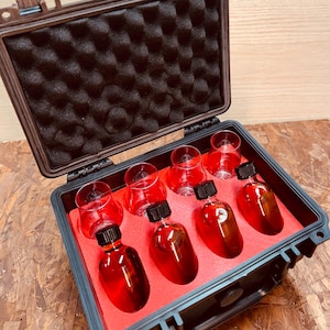 May include: A black, hard-shell case with a red interior holds six small glass bottles filled with a red liquid and six small clear glasses. The case is open, revealing the contents. The bottles have black caps.