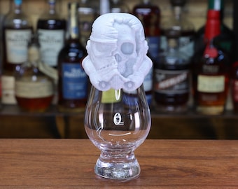 May include: A white glass whisky glass with a white ceramic skull and stormtrooper helmet topper. The glass is sitting on a wooden surface.