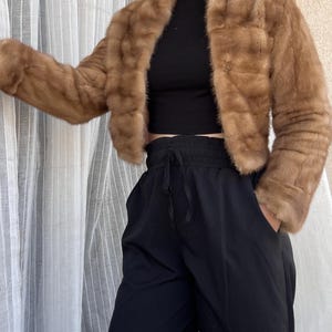 Puede incluir: Una chaqueta de piel marrón con cuello alto y mangas largas. La chaqueta es corta y tiene un corte holgado. La modelo lleva la chaqueta sobre una camiseta negra y pantalones negros.