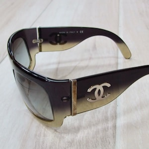 May include: Black and gold gradient sunglasses with a silver Chanel logo on the side. The sunglasses have a thick frame and a wide lens.