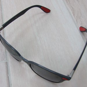 以下が含まれることがあります： 黒いサングラス。レンズは濃い色で、フレームは長方形です。テンプルは黒で、先端に赤いアクセントがあります。モダンなデザインで、アウトドア活動に適しています。