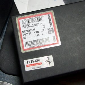 以下が含まれることがあります： 「ORB8351M F60171 *3N ITA 140 A 60」のテキストが記載された赤と白のラベルが付いた黒い箱。小さなラベルには「Ferrari OFFICIAL PRODUCT」と記載されています。箱は暗い表面にあります。