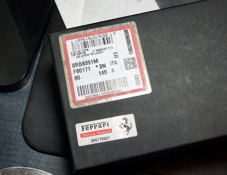 以下が含まれることがあります： 「ORB8351M F60171 *3N ITA 140 A 60」のテキストが記載された赤と白のラベルが付いた黒い箱。小さなラベルには「Ferrari OFFICIAL PRODUCT」と記載されています。箱は暗い表面にあります。