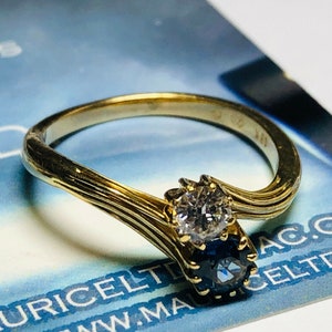 Pu&ograve; includere: Un anello d'oro con un diamante e uno zaffiro. L'anello &egrave; incastonato in una fascia attorcigliata.