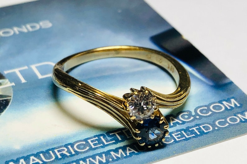 Pu&ograve; includere: Un anello d'oro con un diamante e uno zaffiro. L'anello &egrave; incastonato in una fascia attorcigliata.