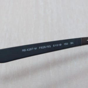 以下が含まれることがあります： 黒いサングラスのクローズアップ。濃い色のレンズと、テンプルに赤いアクセントがあります。サングラスのテンプルには「RB 4297-M F626/SG 51019 150 3N」の文字があります。