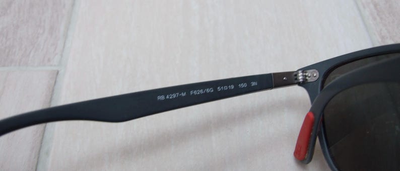 以下が含まれることがあります： 黒いサングラスのクローズアップ。濃い色のレンズと、テンプルに赤いアクセントがあります。サングラスのテンプルには「RB 4297-M F626/SG 51019 150 3N」の文字があります。