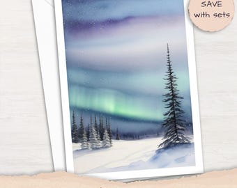 May include: A greeting card featuring a watercolor illustration of a snowy landscape with evergreen trees under a vibrant aurora borealis sky in shades of purple, blue, and green. The card is paired with a white envelope and a circular tag.
