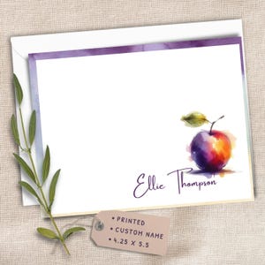 May include: A white stationery card with a purple watercolor border, featuring a colorful watercolor apple illustration and the printed name Ellie Thompson. A small tag attached to the card displays the text: PRINTED, CUSTOM NAME, 4.25 X 5.5.