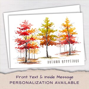 May include: A white greeting card featuring a watercolor illustration of three trees with red, orange, and yellow foliage. The text "autumn greetings" is printed in black in the bottom right corner. The card is placed on a white envelope.