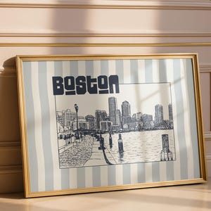 May include: Framed art print featuring a black and white illustration of the Boston skyline. The word "BOSTON" is printed in bold, dark blue letters above the illustration. The artwork is set against a striped background and framed in gold.