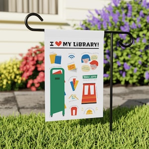 May include: A white garden flag with the text "I ❤️ MY LIBRARY!" and colorful illustrations of books, a library card, and other library-related icons. The flag is displayed on a black metal stand in a grassy yard.