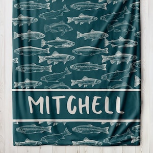 May include: A teal blue fleece blanket with a white pattern of fish and the name "MITCHELL" printed in white.