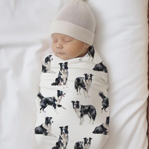 May include: A white swaddle blanket with a black and white border collie print. A baby is sleeping in the swaddle, wearing a white knit hat.