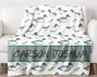 May include: White fleece throw blanket with a green and white watercolor duck pattern. The blanket has a green band with the names "CARSON JOSHUA" written in black.