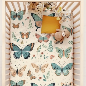 May include: A white crib sheet with a butterfly and floral pattern. The sheet features butterflies in shades of blue, orange, and pink, along with green leaves and small pink flowers.