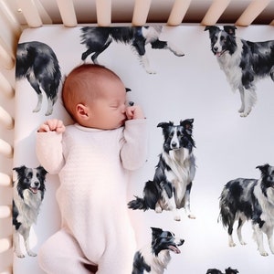May include: A white cot sheet with a black and white border collie dog print. A baby is sleeping on the sheet.