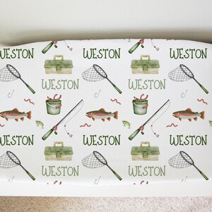 May include: White fabric with a repeating pattern of fishing-themed illustrations. The illustrations include a fishing rod, a bucket of worms, a net, a tackle box, and a trout. The name "WESTON" is repeated throughout the pattern.