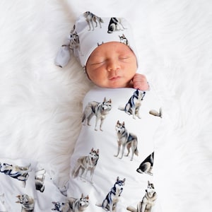 May include: A newborn baby swaddled in a white blanket and matching cap, both adorned with watercolor illustrations of Siberian Huskies. The baby is nestled on a fluffy white surface.