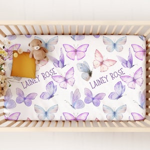 May include: A white cot sheet with a purple and pink butterfly pattern. The sheet has the name "Lainey Rose" printed on it in purple.