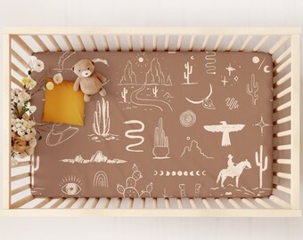 May include: A brown fitted crib sheet with a white desert-themed pattern. The pattern includes cacti, mountains, stars, a crescent moon, a snake, a cowboy riding a horse, and an eagle. A stuffed teddy bear and a yellow pillow are on the sheet.