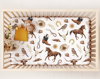 May include: A white crib sheet with a western-themed print featuring horses, cowboy hats, boots, and other western imagery. The sheet is in a crib with a wooden frame.