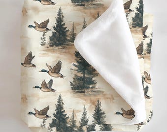 May include: A folded blanket featuring a watercolor design of flying ducks and evergreen trees. The blanket has a cream-colored background with a soft, white, plush interior. The ducks are brown with blue and green accents.
