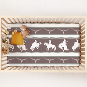 May include: A white crib with a brown and white striped fitted sheet. The sheet features a repeating pattern of cowboys riding horses and steer skulls. A stuffed animal is on the sheet.