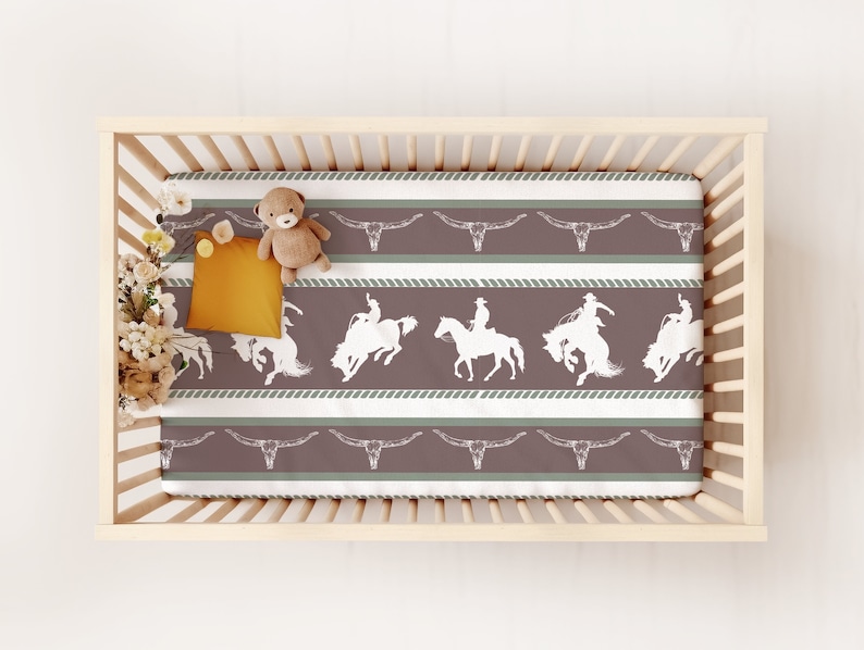 May include: A white crib with a brown and white striped fitted sheet. The sheet features a repeating pattern of cowboys riding horses and steer skulls. A stuffed animal is on the sheet.