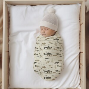 May include: A newborn baby swaddled in a white and green swaddle with a fish and plant pattern. The baby is wearing a white knit hat with a pom pom on top. The baby is lying on a white sheet in a wooden crib.