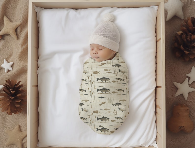 May include: A newborn baby swaddled in a white and green swaddle with a fish and plant pattern. The baby is wearing a white knit hat with a pom pom on top. The baby is lying on a white sheet in a wooden crib.
