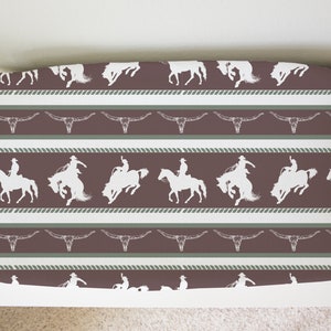 May include: A brown and white fabric with a repeating pattern of cowboys riding horses and steer skulls. The fabric has a rope-like border.