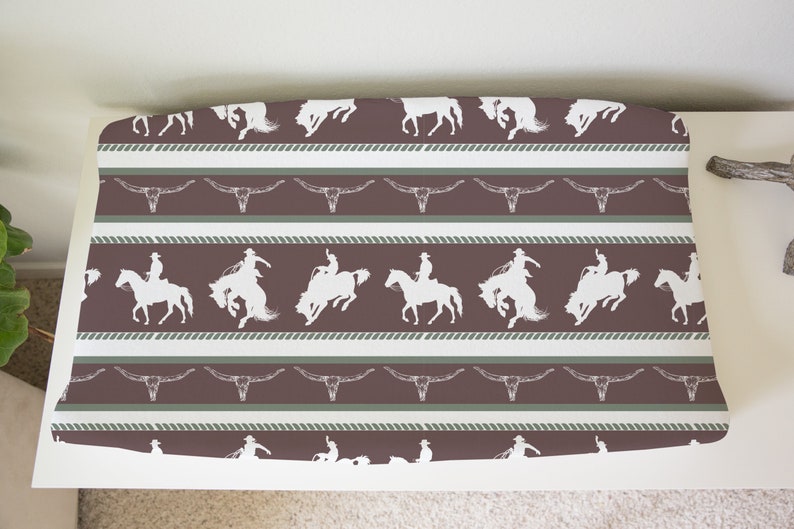 May include: A brown and white fabric with a repeating pattern of cowboys riding horses and steer skulls. The fabric has a rope-like border.