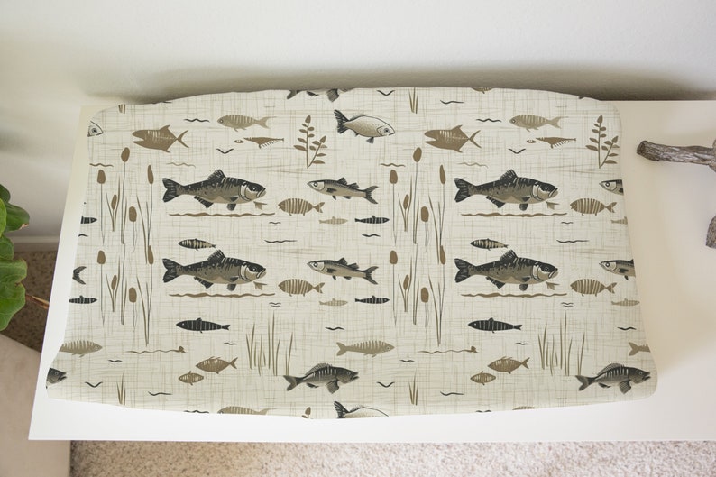 May include: A beige fabric with a repeating pattern of brown fish and reeds. The fish are stylized and have a dark brown outline. The reeds are a lighter brown and have a more natural look.