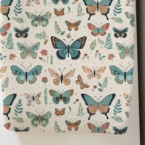 May include: A cream-colored fabric with a repeating pattern of colorful butterflies. The butterflies are in shades of blue, green, orange, and pink. The fabric has a subtle texture.