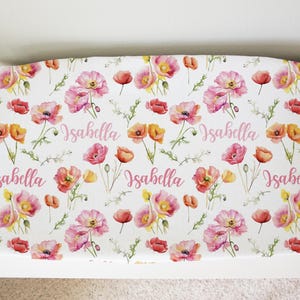 May include: A white changing pad cover with a pink poppy floral pattern and the name "Isabella" repeated four times in pink.