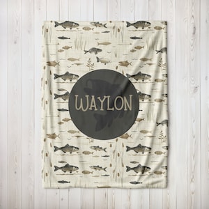 May include: A cream-colored blanket with a pattern of brown fish and reeds. A large black circle with the name "WAYLON" in white text is centered on the blanket.