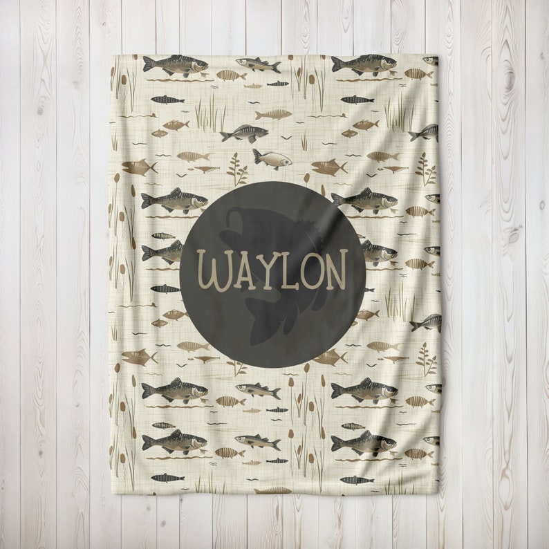 May include: A cream-colored blanket with a pattern of brown fish and reeds. A large black circle with the name "WAYLON" in white text is centered on the blanket.