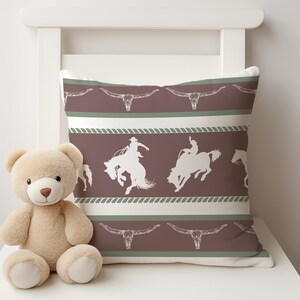 May include: A brown and white throw pillow with a repeating pattern of cowboys riding horses and steer skulls. The pillow is on a white chair with a tan teddy bear in front of it.