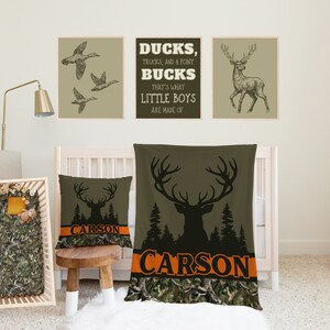 May include: A nursery room with a cot, a blanket, and a pillow featuring a camouflage pattern and the name "CARSON" printed on it. There are three framed prints on the wall with images of ducks, a deer, and the text "DUCKS, TRUCKS, AND A POINT BUCKS THAT'S WHAT LITTLE BOYS ARE MADE OF".