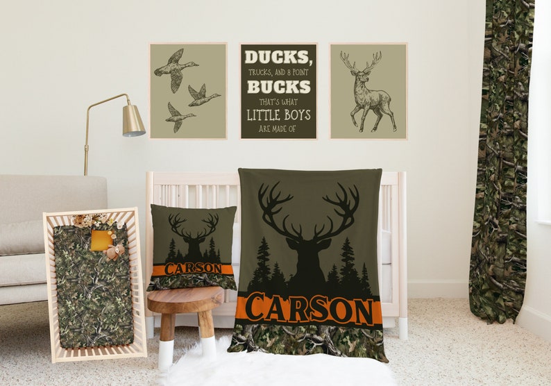 May include: A nursery room with a cot, a blanket, and a pillow featuring a camouflage pattern and the name "CARSON" printed on it. There are three framed prints on the wall with images of ducks, a deer, and the text "DUCKS, TRUCKS, AND A POINT BUCKS THAT'S WHAT LITTLE BOYS ARE MADE OF".