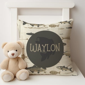 May include: A beige pillow with a brown and black fish pattern. The pillow has a large black circle with the name "WAYLON" in white text.