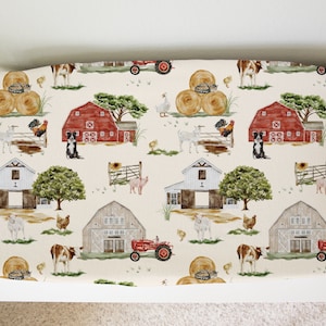 May include: A cream-colored fabric with a repeating pattern of farm animals, barns, hay bales, and tractors. The pattern includes red and white barns, brown hay bales, white goats, brown cows, red tractors, and black and white border collies.