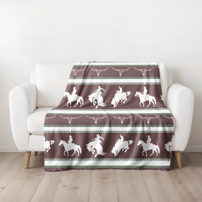 May include: A brown and green fleece throw blanket with a repeating pattern of white silhouettes of cowboys riding horses. The pattern includes a rope design and a skull motif.